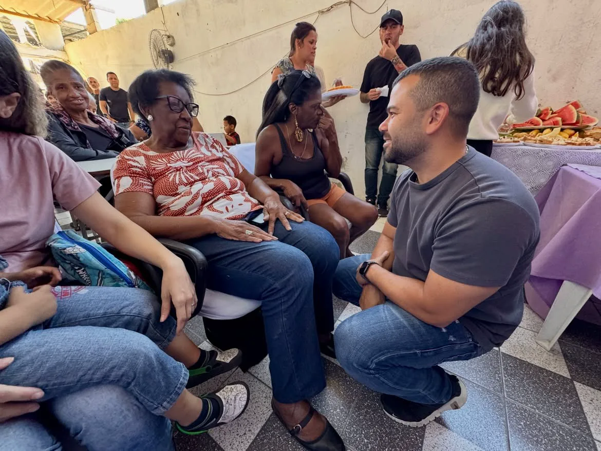 Felipe Pampolha agachado ao lado de moradoras sentadas em cadeiras, ouvindo uma senhora falar durante um encontro comunitário.