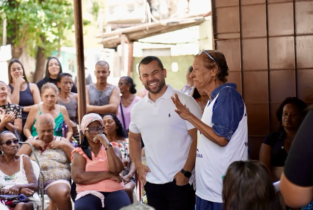 Felipe Pampolha rindo ao lado de uma senhora que gesticula enquanto fala, diante de uma plateia de moradoras sentadas.